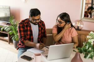 millennial indian husband and wife doing domestic paperwork, accounting job and reviewing paper bills, receipts at laptop computer, using online calculator - economic crisis concept photo