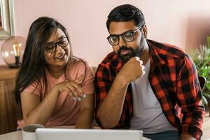 Indian family couple cuddle at desk make video call to friends using laptop webcam. Loving young spouses look at computer screen waving hands in good mood greeting parents communicating online via app photo