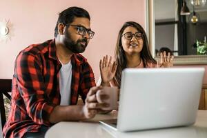 Indian family couple cuddle at desk make video call to friends using laptop webcam. Loving young spouses look at computer screen waving hands in good mood greeting parents communicating online via app photo