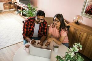 Indian family couple cuddle at desk make video call to friends using laptop webcam. Loving young spouses look at computer screen waving hands  communicating online via app photo