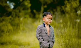 linda pequeño chico un bien vestido chico en un traje en un patio interior con un césped y mirando para alguna cosa interesante. foto