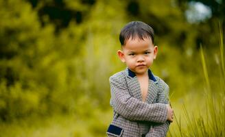 linda pequeño chico un bien vestido chico en un traje en un patio interior con un césped y mirando para alguna cosa interesante. foto