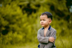 linda pequeño chico un bien vestido chico en un traje en un patio interior con un césped y mirando para alguna cosa interesante. foto