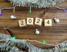 New Year's card 2024. Wooden cubes with numbers 2024, tinsel, garland and New Year's balls on a wooden background. photo