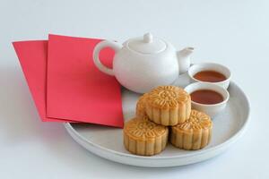 Chinese Mid-Autumn Festival concept. Mooncakes and hot tea pot and red envelopes in plate isolated on white background. Chinese mid autumn festival food. photo