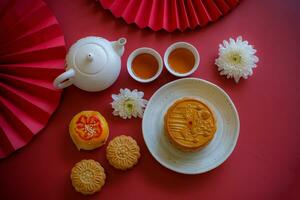 Chinese Mid-Autumn Festival concept made from mooncakes, tea decorated with Chrysanthemum blossom, rabbit and red paper fans on red background. photo
