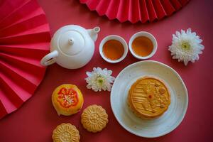 Chinese Mid-Autumn Festival concept made from mooncakes, tea decorated with Chrysanthemum blossom, rabbit and red paper fans on red background. photo