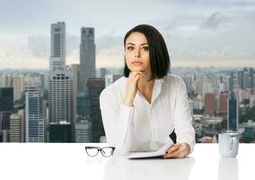Pensive young business woman in white shirt on her desk with paper documents over city background. Achievement business career concept. photo