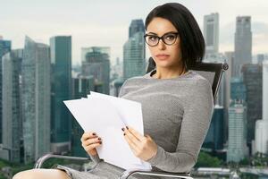 Pensive young business woman in glasses sitting on chair with paper documents over Singapore city background. .Achievement business career concept. photo