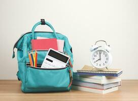 Front view  of green backpack with school supplie, stack of books and white vintage alarm clock 8 o'clock on wooden table and white  background with copy space. photo