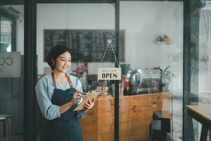 Beautiful young barista woman in apron holding order paper and standing in front of the door of cafe with open sign board. Business owner startup concept. photo