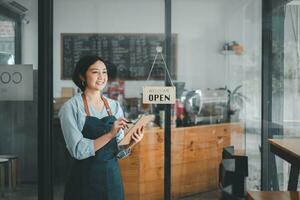 Beautiful young barista woman in apron holding order paper and standing in front of the door of cafe with open sign board. Business owner startup concept. photo