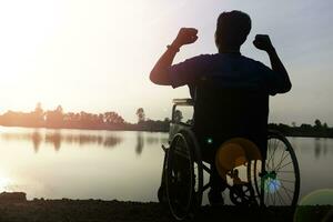 Silhouette young disabled man seeing river background.He is raise a hand to and sitting on wheelchair.despair,lonely,hope.Photo concept depression and Patient. photo