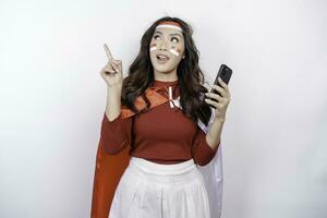 A happy Asian woman wearing headband, holding her phone, and pointing copy space on top of her, isolated by white background. Indonesia's independence day photo