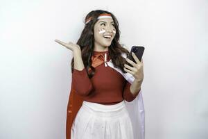 A happy Asian woman wearing headband, holding her phone, and pointing copy space beside her, isolated by white background. Indonesia's independence day photo