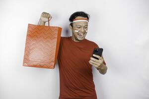 An attractive Asian man standing excited holding an online shopping bag and his smartphone, studio shot isolated on white background. Indonesia's Independence day concept photo