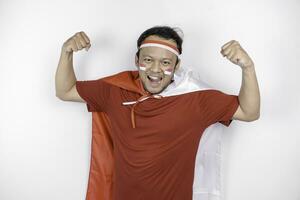 Excited Asian man wearing a red top, flag cape and headband, showing strong gesture by lifting his arms and muscles smiling proudly. Indonesia's independence day concept. photo