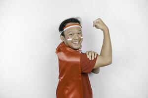 Excited Asian man wearing a red top, flag cape and headband, showing strong gesture by lifting his arms and muscles smiling proudly. Indonesia's independence day concept. photo