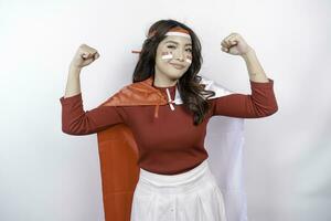 Excited Asian woman wearing a red top, flag cape and headband, showing strong gesture by lifting her arms and muscles smiling proudly. Indonesia's independence day concept. photo