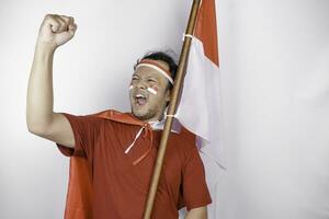 A young Asian man with a happy successful expression wearing red top and headband while holding Indonesia's flag, isolated by white background. Indonesia's independence day concept. photo