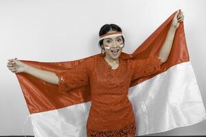 Happy smiling Indonesian woman wearing red kebaya and headband holding Indonesia's flag to celebrate Indonesia Independence Day isolated over white background. photo