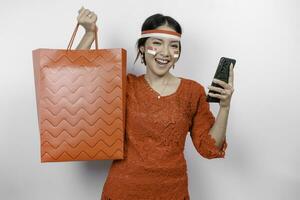 An excited Asian woman wearing red kebaya holding a shopping bag and her smartphone, studio shot isolated on white background. Indonesia's Independence day concept photo