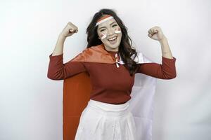 Excited Asian woman wearing a red top, flag cape and headband, showing strong gesture by lifting her arms and muscles smiling proudly. Indonesia's independence day concept. photo