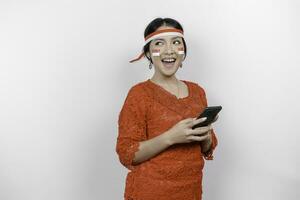 A portrait of a smiling Asian woman wearing red kebaya and headband and holding her phone, isolated by white background. Indonesia's independence day concept photo