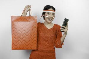 An excited Asian woman wearing red kebaya holding a shopping bag and her smartphone, studio shot isolated on white background. Indonesia's Independence day concept photo