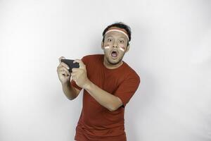 A portrait of a shocked Asian man wearing headband, holding his phone while his mouth wide open, isolated by white background. Indonesia's independence day concept photo