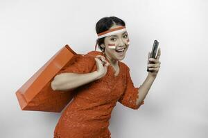 An excited Asian woman wearing red kebaya holding a shopping bag and her smartphone, studio shot isolated on white background. Indonesia's Independence day concept photo