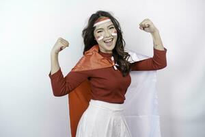 Excited Asian woman wearing a red top, flag cape and headband, showing strong gesture by lifting her arms and muscles smiling proudly. Indonesia's independence day concept. photo