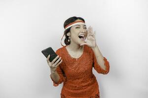 Young beautiful woman wearing a red kebaya is holding her phone while shouting and screaming loud with a hand on her mouth. Indonesia's independence day concept. photo