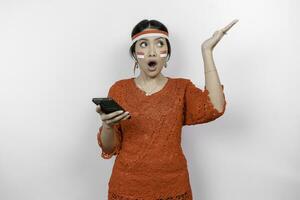 A portrait of a shocked Asian woman wearing red kebaya and headband, holding her phone, and pointing copy space on top of her, isolated by white background. Indonesia's independence day concept photo