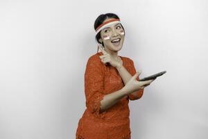 A happy Asian woman wearing red kebaya and headband, holding her phone, and pointing copy space on top of her, isolated by white background. Indonesia's independence day photo