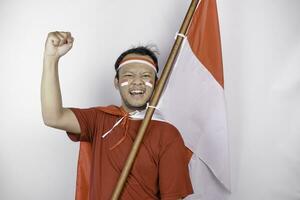 un joven asiático hombre con un contento exitoso expresión vistiendo rojo parte superior y venda mientras participación de indonesia bandera, aislado por blanco antecedentes. de indonesia independencia día concepto. foto