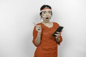 A portrait of a shocked Asian woman wearing red kebaya and headband, holding her phone, and pointing copy space on top of her, isolated by white background. Indonesia's independence day concept photo