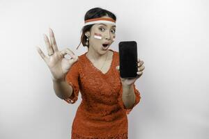 A cheerful Asian woman wearing red kebaya and headband, showing her phone while gesturing OK sign with her fingers, isolated by white background. Indonesia's independence day concept photo
