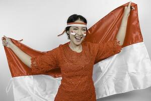 Happy smiling Indonesian woman wearing red kebaya and headband holding Indonesia's flag to celebrate Indonesia Independence Day isolated over white background. photo