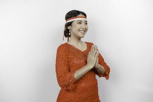 A friendly Indonesian woman is wearing red kebaya gesturing traditional greeting and Indonesia's flag headband to celebrate Indonesia Independence Day. Isolated by white background. photo