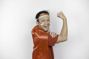 Excited Asian man wearing a red top, flag cape and headband, showing strong gesture by lifting his arms and muscles smiling proudly. Indonesia's independence day concept. photo