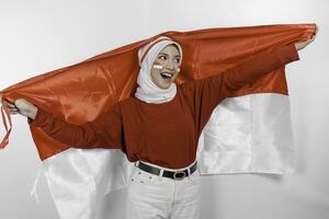 Happy smiling Indonesian muslim woman wearing red top and white hijab holding Indonesia's flag to celebrate Indonesia Independence Day. Isolated by white background. photo