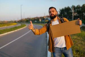 Man is hitchhiking on roadside trying to stop car. He is holding blank cardboard for your text. photo