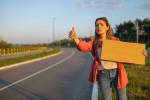 Woman is hitchhiking on roadside trying to stop car. She is holding blank cardboard for your text. photo