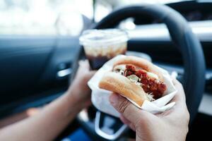 asiático mujer conductor sostener y comer hamburguesa en auto, peligroso y riesgo un accidente. foto