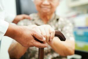 Doctor help Asian elderly disability woman patient holding walking stick in wrinkled hand at hospital. photo