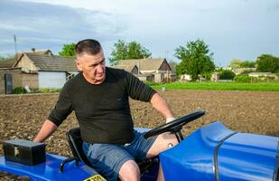 Kherson oblast, Ukraine - May 29, 2021 An elderly farmer sits and drives a tractor. Seasonal worker. Recruiting and hiring employees for work in farm. Milling soil. Loosening , cultivation. Plowing photo