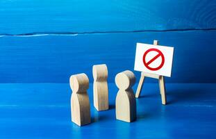 A group of people figurines looking at an easel with a red prohibitory symbol NO. Expression of protest and disagreement. Inaccessibility and absence. Quarantine and lockdown. Hard restrictions photo