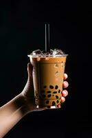 A hand holding takeaway plastic cup of delicious iced bubble milk tea and black pearls isolated on black background. ai generated photo