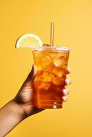 A hand holding takeaway plastic cup of delicious iced lemon tea isolated on yellow background. ai generated photo